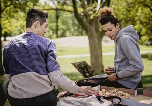 two guys eating pizza in a park with a laptop by them